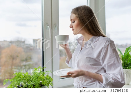Portrait of young woman with cup of coffee near winter autumn window Portrait of young woman with cup of coffee near winter autumn window 81764138