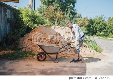 A 30-year-old man pours sand from a large pile of sand onto a garden wheelbarrow A 30-year-old man pours sand from a large pile of sand onto a garden wheelbarrow 81765340