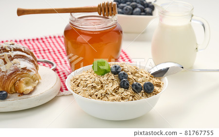 Morning breakfast, raw oatmeal flakes in a ceramic plate, milk in a decanter, blueberries and honey in a jar on a white table Morning breakfast, raw oatmeal flakes in a ceramic plate, milk in a decanter, blueberries and honey in a jar on a white table 81767755