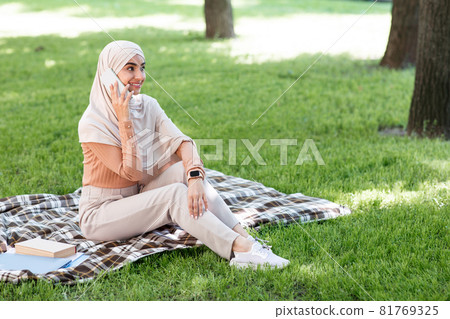Smiling young middle eastern woman in hijab calling by phone, relax in park and enjoying sunny day Smiling young middle eastern woman in hijab calling by phone, relax in park and enjoying sunny day 81769325