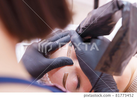 The master applies eyebrow makeup paint to the client with a tattoo machine. 81773452