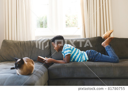 Happy african american boy reading book and lying on couch with cat in living room 81780755