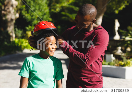 African american father with son smiling and putting on helmet before skateboarding in garden 81780889