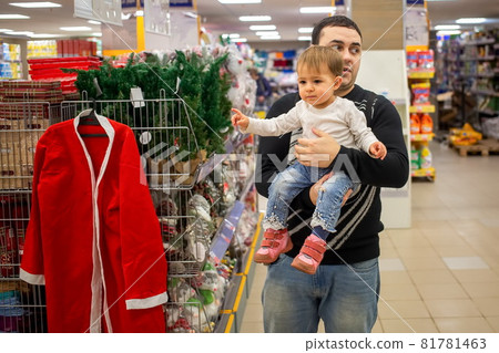 A young Caucasian man holds a small cute baby in his arms in a store near a bookcase. baby points with finger at Christmas Santa costume and New Year decorations. close-up, soft focus, blur background A young Caucasian man holds a small cute baby in his arms in a store near a bookcase. baby points with finger at Christmas Santa costume and New Year decorations. close-up, soft focus, blur background 81781463