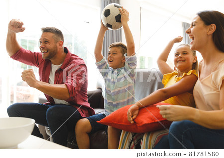 Excited caucasian parents on couch with daughter and son watching football match on tv and cheering 81781580