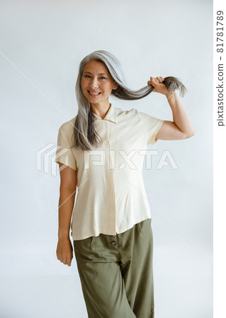Positive Asian lady holds lock of healthy silver hair on light grey background 81781789