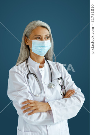 Hoary haired Asian woman doctor with mask and crossed arms stands on blue background 81781810
