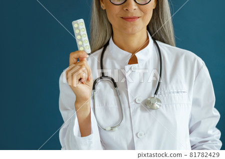 Happy mature lady therapist in white coat shows pills on blue background Happy mature lady therapist in white coat shows pills on blue background 81782429