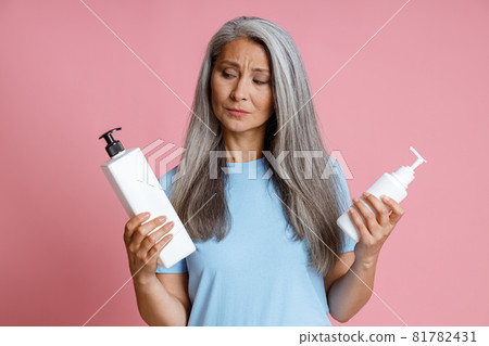 Thoughtful middle aged Asian lady chooses cosmetic product on pink background 81782431