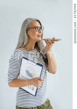 Mature Asian businesswoman records audio message on cellphone holding diagrams in studio 81782444