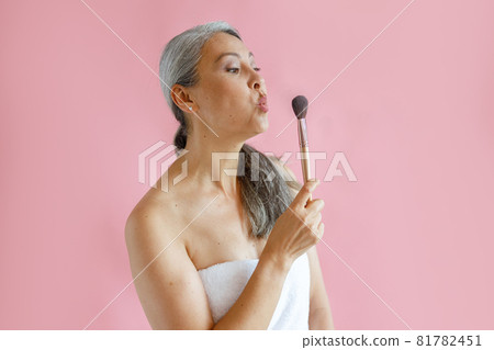 Funny middle aged Asian lady wrapped with white towel blows cosmetic brush on pink background Funny middle aged Asian lady wrapped with white towel blows cosmetic brush on pink background 81782451