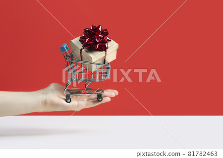 Hand holding Toy shopping cart or trolley with large gift box. A gift for a holiday, anniversary, or birthday. Trolley cart with a gift box Isolated over red background. Banner with copy space 81782463