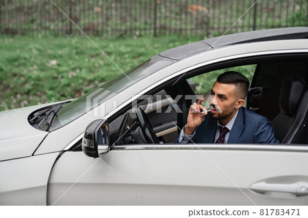 a man sits behind the wheel of a car with the door open and smokes a cigarette. a man sits behind the wheel of a car with the door open and smokes a cigarette. 81783471