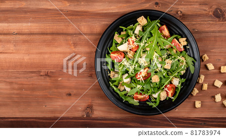 Rocket salad with tomato, crispy crouton, small pieces of herb roasted chicken and parmesan cheese in black dish appear on right side of picture decorated with extra crouton on the wooded table Rocket salad with tomato, crispy crouton, small pieces of herb roasted chicken and parmesan cheese in black dish appear on right side of picture decorated with extra crouton on the wooded table 81783974