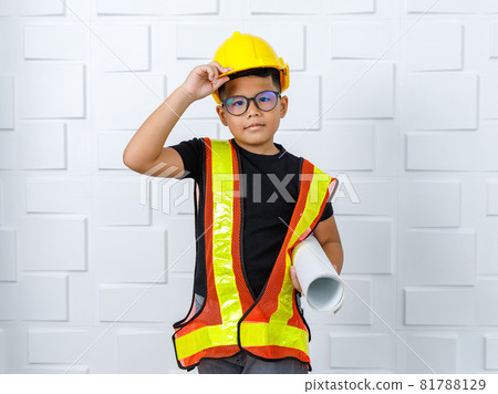 Young Asian boy wearing glasses, black shirt, yellow safety vest and protective helmet as engineer, happily holding paper roll and standing with smile near white wall as enjoy construction work 81788129