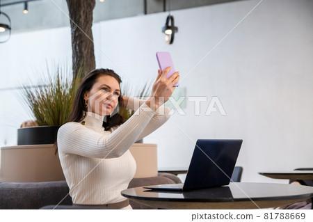 beautiful caucasian woman makes selfie on a smartphone sitting at a table with a laptop. a blogger creates content, a blog author works in a cafe or coworking space. beautiful caucasian woman makes selfie on a smartphone sitting at a table with a laptop. a blogger creates content, a blog author works in a cafe or coworking space. 81788669