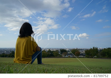 一個坐在後面的女人坐在藍天下的草地上，在公園裡，可以看到風景 81789752