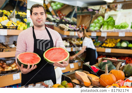 Young man seller holding half of watermelon in hands in fruit store Young man seller holding half of watermelon in hands in fruit store 81792174