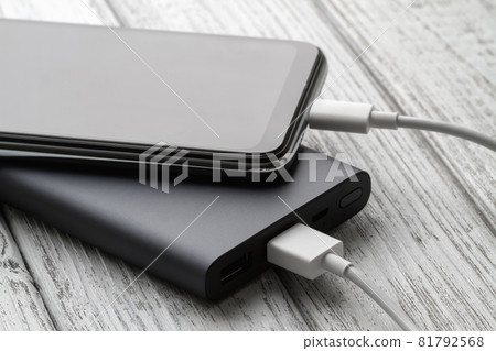 The smartphone is charging from external powerbank with cable on a gray wooden background. Closeup, selective focus The smartphone is charging from external powerbank with cable on a gray wooden background. Closeup, selective focus 81792568