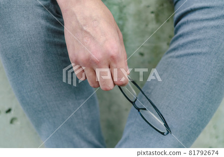 A close-up of a man's hand holding his glasses. Businessman with gray pants. Outside the room. Selective focus. 81792674