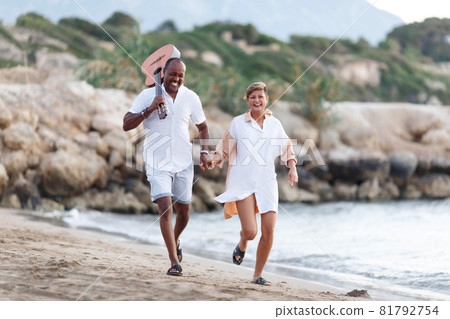 Happy mixed race, middle age couple embracing while walking on the beach holding a guitar. Man holding guitar with woman. 81792754
