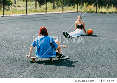 A young girl is sitting on a skateboard outdoors on a basketball court with her basketball player friend A young girl is sitting on a skateboard outdoors on a basketball court with her basketball player friend 81793376