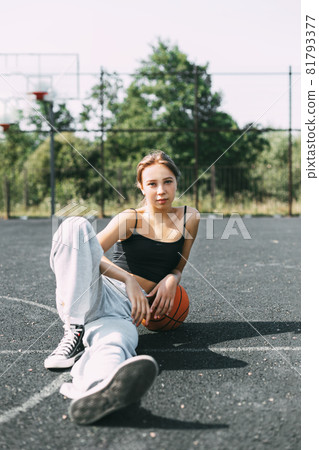 Portrait of a charming girl sitting on a sports field in a park or school with a basketball after a game or workout Portrait of a charming girl sitting on a sports field in a park or school with a basketball after a game or workout 81793377