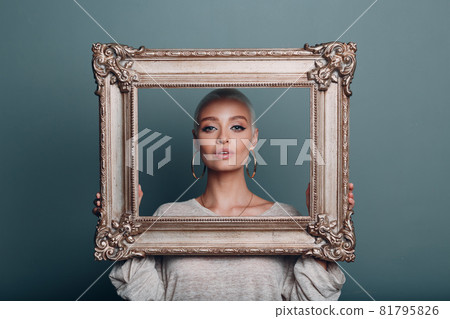Millenial young woman with short blonde hair holds gilded picture frame in hands behind her face portrait. 81795826