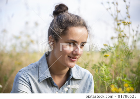 beautiful young woman sits in a meadow in flowers on a sunny summer day and smiles. beautiful young woman sits in a meadow in flowers on a sunny summer day and smiles. 81796523