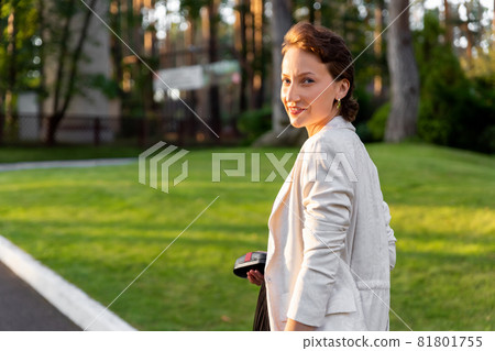 Portrait of beautiful young adult elegant stylish caucasian woman walking by green garden park in warm evening golden sunset rays outdoors. Confident happy smiling female person enjoy walk outside 81801755