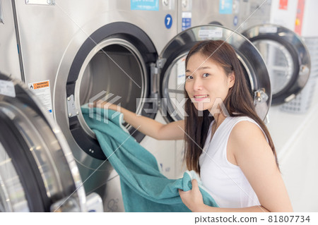 In the self-service laundry with dryer machines in the backdrop, a young woman enjoys clean ironed garments. In the self-service laundry with dryer machines in the backdrop, a young woman enjoys clean ironed garments. 81807734