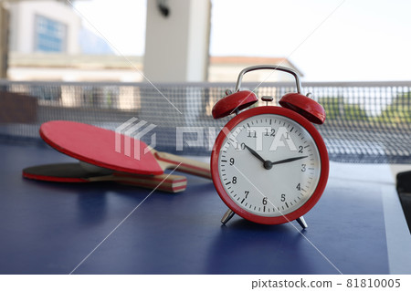 Red alarm clock standing on tennis table near rackets closeup 81810005