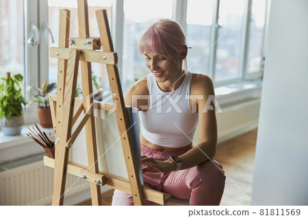Smiling woman with pink hair draws on canvas at wooden easel on balcony 81811569
