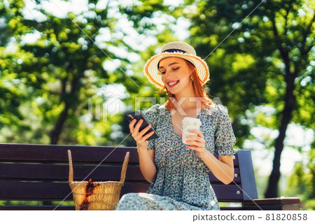 Beautiful redhead young woman relaxing on the bench at the city park, holding coffee in hands, using phone. Wearing in dress straw hat fashionable earrings 81820858