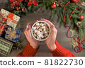 A woman holds a hot cup cocoa with marshmallows. Christmas decorations, gifts and wooden background.  81822730