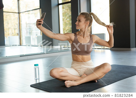 Portrait of young sportive happy girl, coach in sportswear taking selfie before yoga class on sports mat at meditation center, indoors. Concept of healthy lifestyle Portrait of young sportive happy girl, coach in sportswear taking selfie before yoga class on sports mat at meditation center, indoors. Concept of healthy lifestyle 81823284