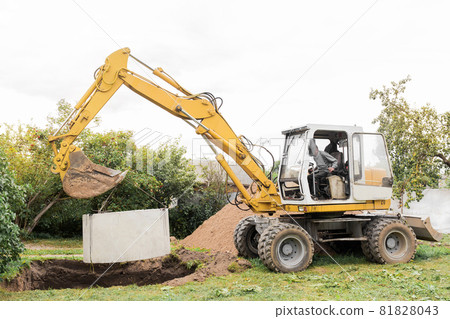 Hydraulic piston system of the excavator with a bucket, lowering into the pit on steel cables concrete sewer ring. Construction or repair of sewer home Hydraulic piston system of the excavator with a bucket, lowering into the pit on steel cables concrete sewer ring. Construction or repair of sewer home 81828043