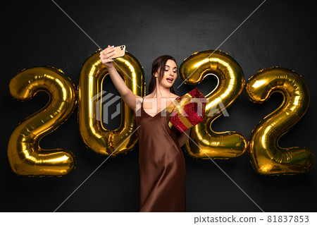 Smiling young brunette woman in elegant dress doing selfie on smartphone on black background golden numbers air balloons. Looking on the red gift box in hand. Happy New Year 2022 celebration holiday  81837853