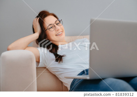 Young woman is resting at home on a white sofa watching a movie on a laptop. 81839312