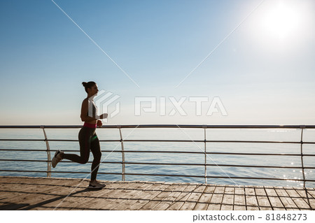 Side view of young fitness woman running along pier. Female athlete workout and jogging on the seaside promenade Side view of young fitness woman running along pier. Female athlete workout and jogging on the seaside promenade 81848273