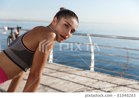 Close-up of attractive sportswoman taking a breath during workout, looking at camera and panting, jogging along seaside promenade 81848283
