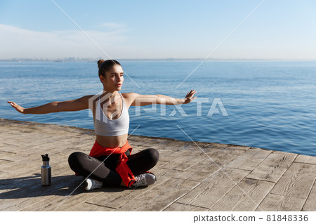 Outdoor shot of relaxed young woman sitting on wooden pier and looking at the sea, doing yoga exercises Outdoor shot of relaxed young woman sitting on wooden pier and looking at the sea, doing yoga exercises 81848336