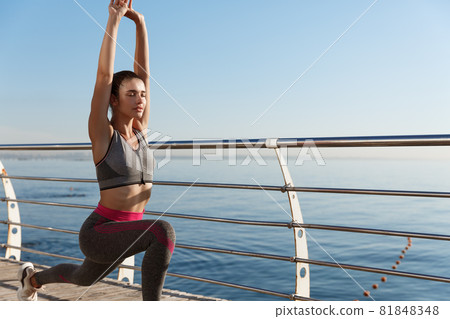 Outdoor shot of sportswoman workout near the sea. Woman workout or doing yoga exercises at the seaside promenade Outdoor shot of sportswoman workout near the sea. Woman workout or doing yoga exercises at the seaside promenade 81848348