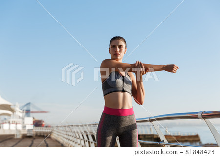Image of beautiful healthy woman in fitness clothing stretching her body before jogging training. Female runner standing on the seaside promenade, training near the sea Image of beautiful healthy woman in fitness clothing stretching her body before jogging training. Female runner standing on the seaside promenade, training near the sea 81848423