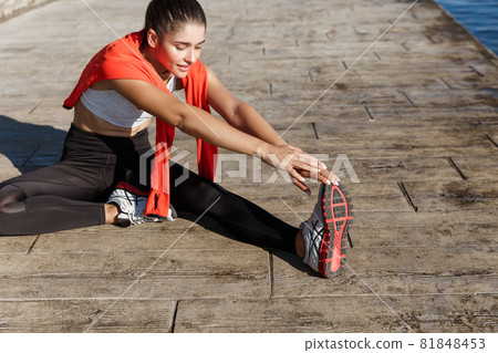 Outdoor shot of fit and healthy sportswoman stretching her legs and workout along seaside promenade 81848453
