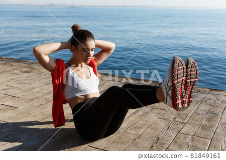 Outdoor shot of confident fitness woman working on her abs. Sportswoman doing crunches with raised legs, training near sea on a pier Outdoor shot of confident fitness woman working on her abs. Sportswoman doing crunches with raised legs, training near sea on a pier 81849161