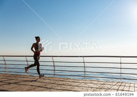 Side view of young fitness woman running along pier. Female athlete workout and jogging on the seaside promenade Side view of young fitness woman running along pier. Female athlete workout and jogging on the seaside promenade 81849194