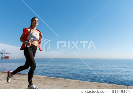 Outdoor shot of smiling sportswoman running along seaside and enjoying the view. Female athlete jogging near the sea 81849304