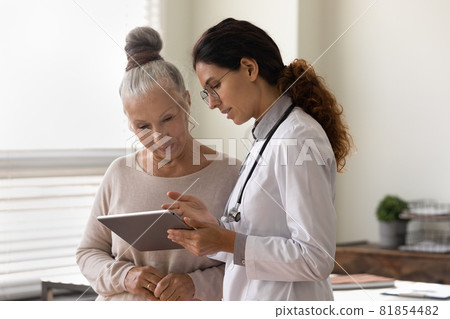 Serious GP doctor showing tablet screen to old female patient 81854482