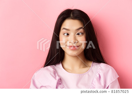 Close-up portrait of funny asian woman squinting eyes and making silly faces, standing against pink background Close-up portrait of funny asian woman squinting eyes and making silly faces, standing against pink background 81857025